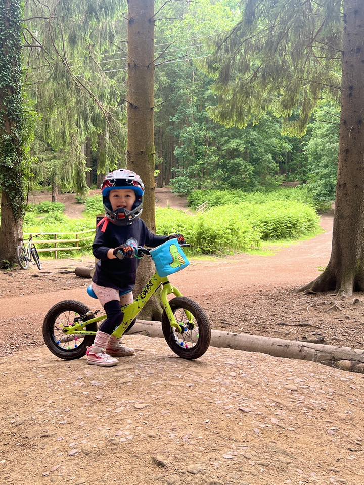 A little girl on a yellow balance bike and wearing a full face helmet, smiling among some trees