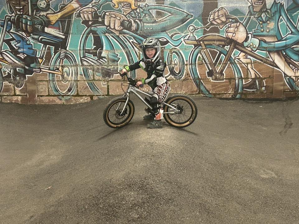 A very young girl standing on a pump track in front of a graffiti wall, over a small pedal bike, wearing a full face helmet and smiling