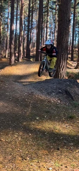 A small child on a small pedal bike, wearing a full face helmet and pictured in mid-air while riding some jumps on a gravel pumptrack