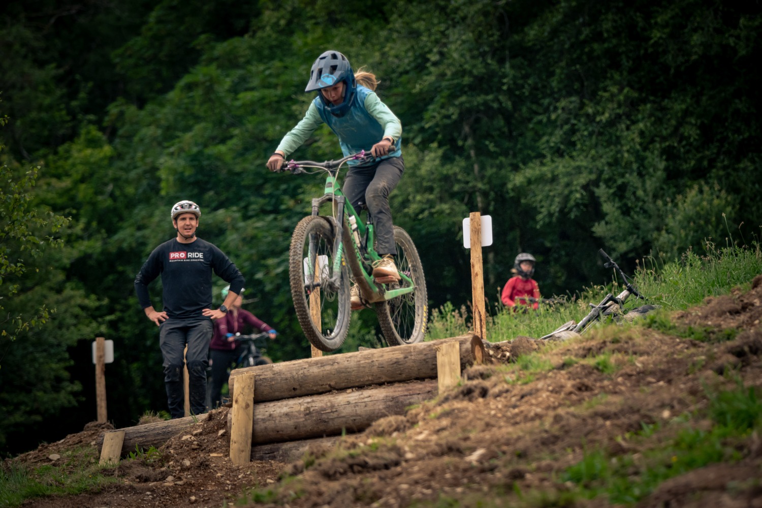 A young girl wearing a full face helmet, about to ride her mountain bike over a large drop off, with a man watching in the background