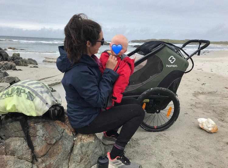A family review of the hamax outback double trailer- a mum holding her baby in a red onesie on the beach with the trailer behind them