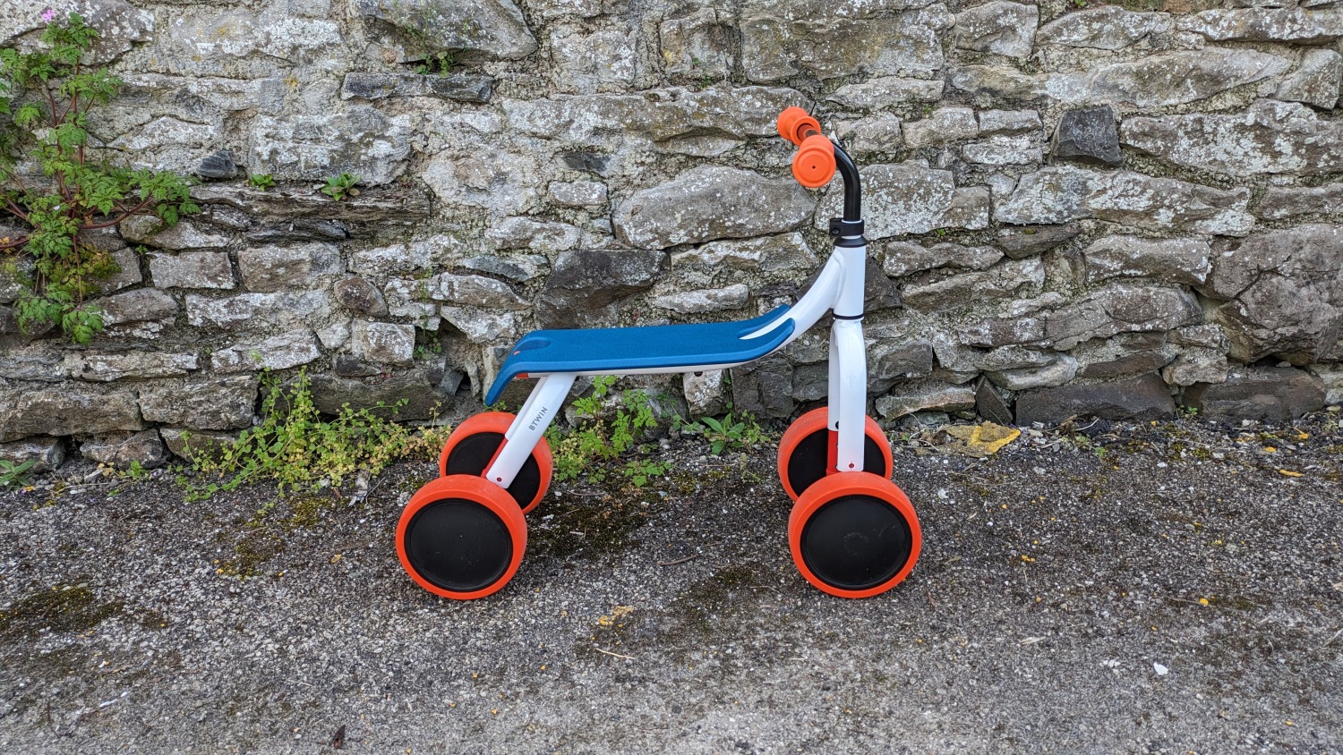 Btwin Convertible 2-in-1 Ride-On to Balance Bike, a white, orange and blue bike leant against a wall