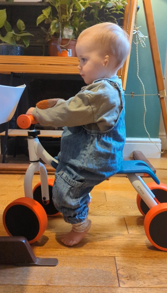 A little girl in dungarees sat on the Btwin Convertible 2-in-1 Ride-On to Balance Bike, a white, orange and blue bike in her living room