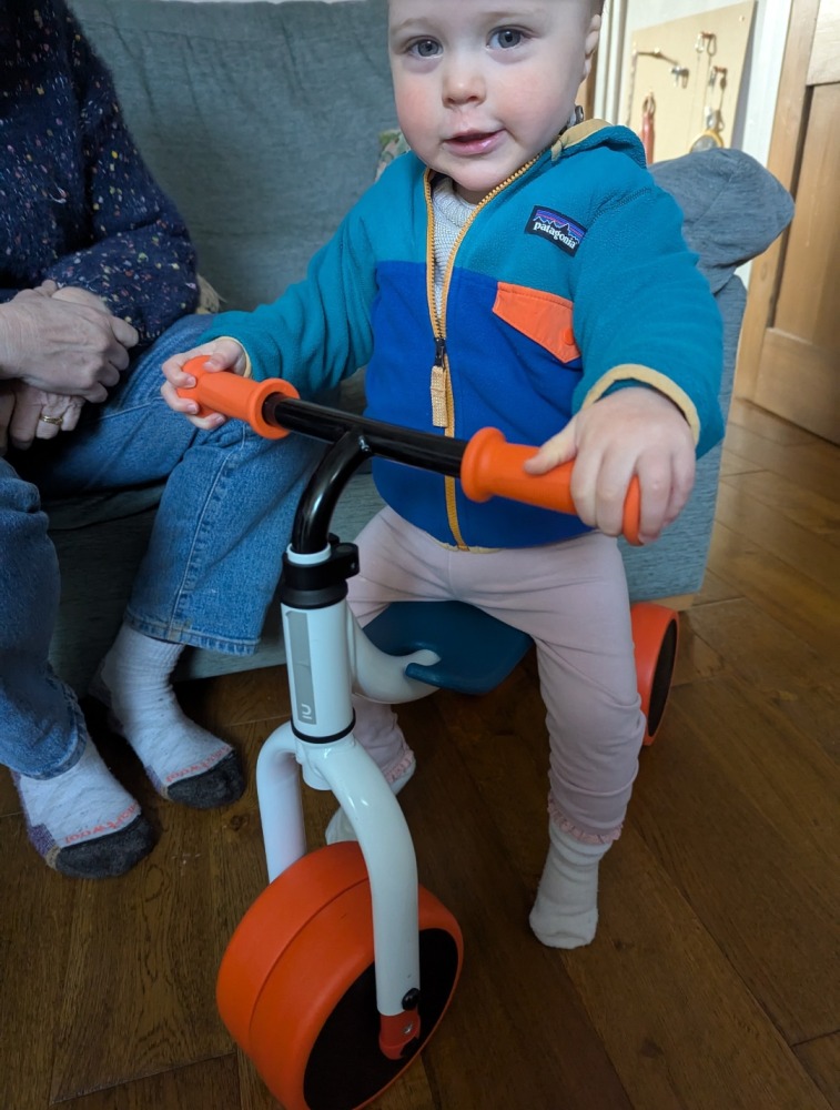 A little girl in a colourful fleece sat on the Btwin Convertible 2-in-1 Ride-On to Balance Bike, a white, orange and blue bike in her living room