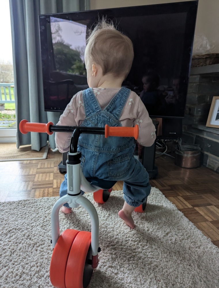 A little girl in dungarees sat backwards on the Btwin Convertible 2-in-1 Ride-On to Balance Bike, a white, orange and blue bike in her living room