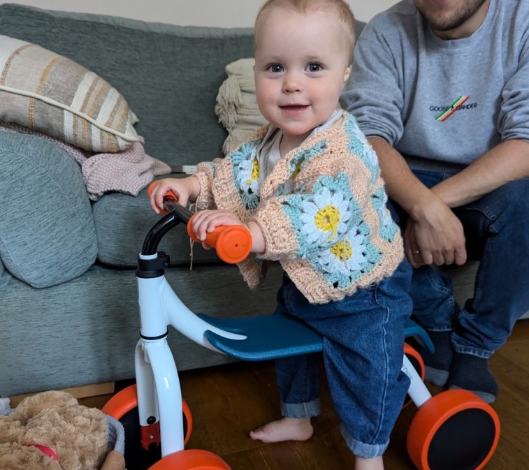 A little girl in a flowery cardigan sat on the Decathlon 2-in-1 ride-on baby balance, a white, orange and blue bike in her living room