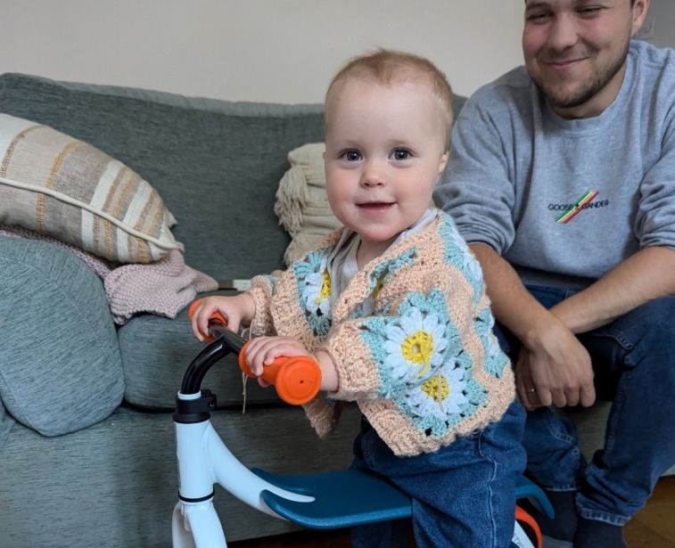 A little girl in a flowery cardigan sat on the Btwin Convertible 2-in-1 Ride-On to Balance Bike, a white, orange and blue bike in her living room