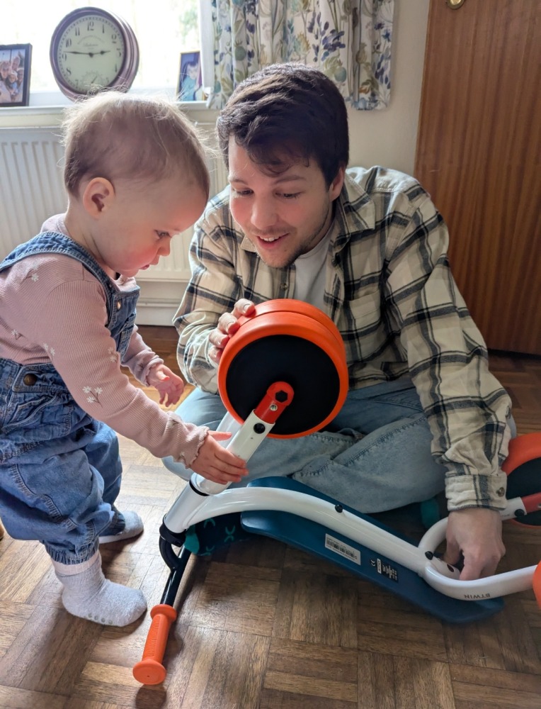A little girl in dungarees is looking at the wheels on the Btwin Convertible 2-in-1 Ride-On to Balance Bike, a white, orange and blue bike with her dad