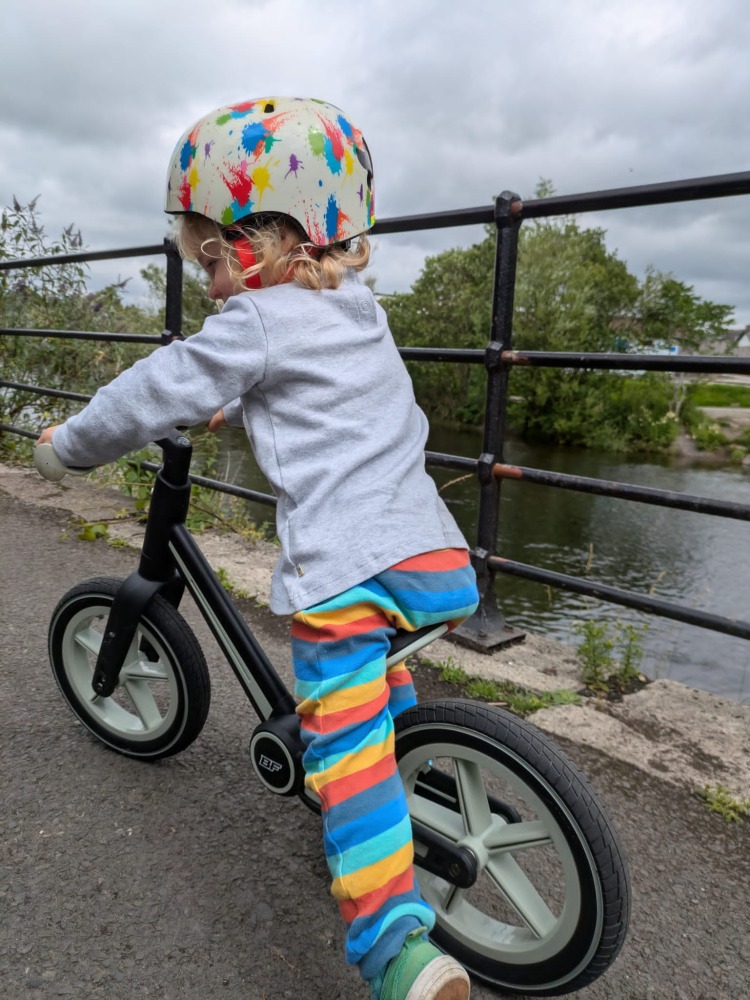 A little boy wearing stripy leggins and a coulourful helmet riding the blue and black Be Fun wheels folding balance bike