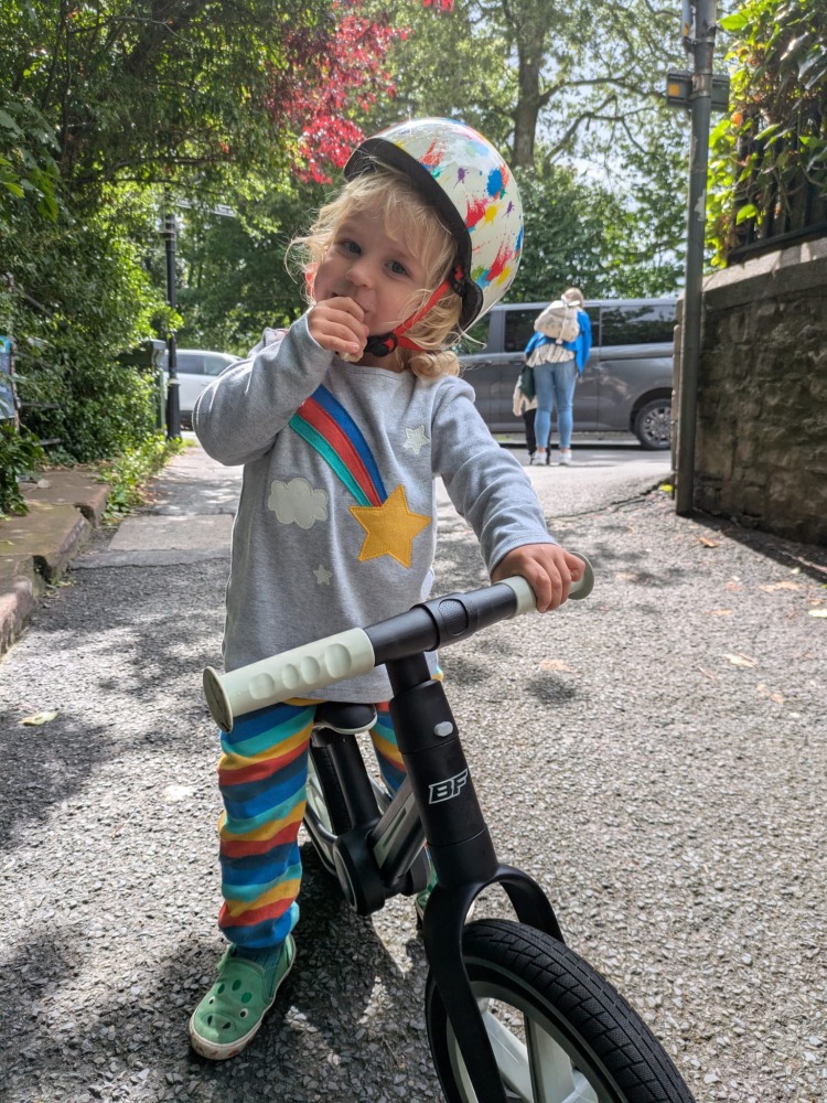 A little boy wearing stripy leggins and a coulourful helmet riding the blue and black Be Fun wheels folding balance bike, stopped to have a snack