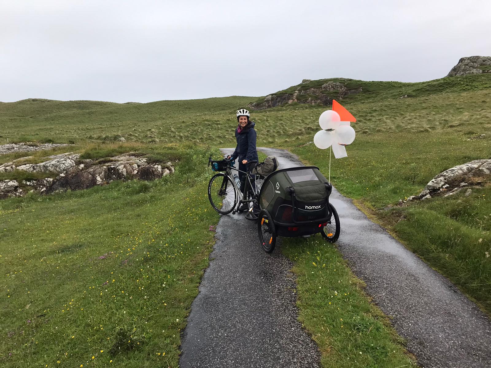 a mum riding on a grassy lane with the Hamax Outback trailer with baloons on the back