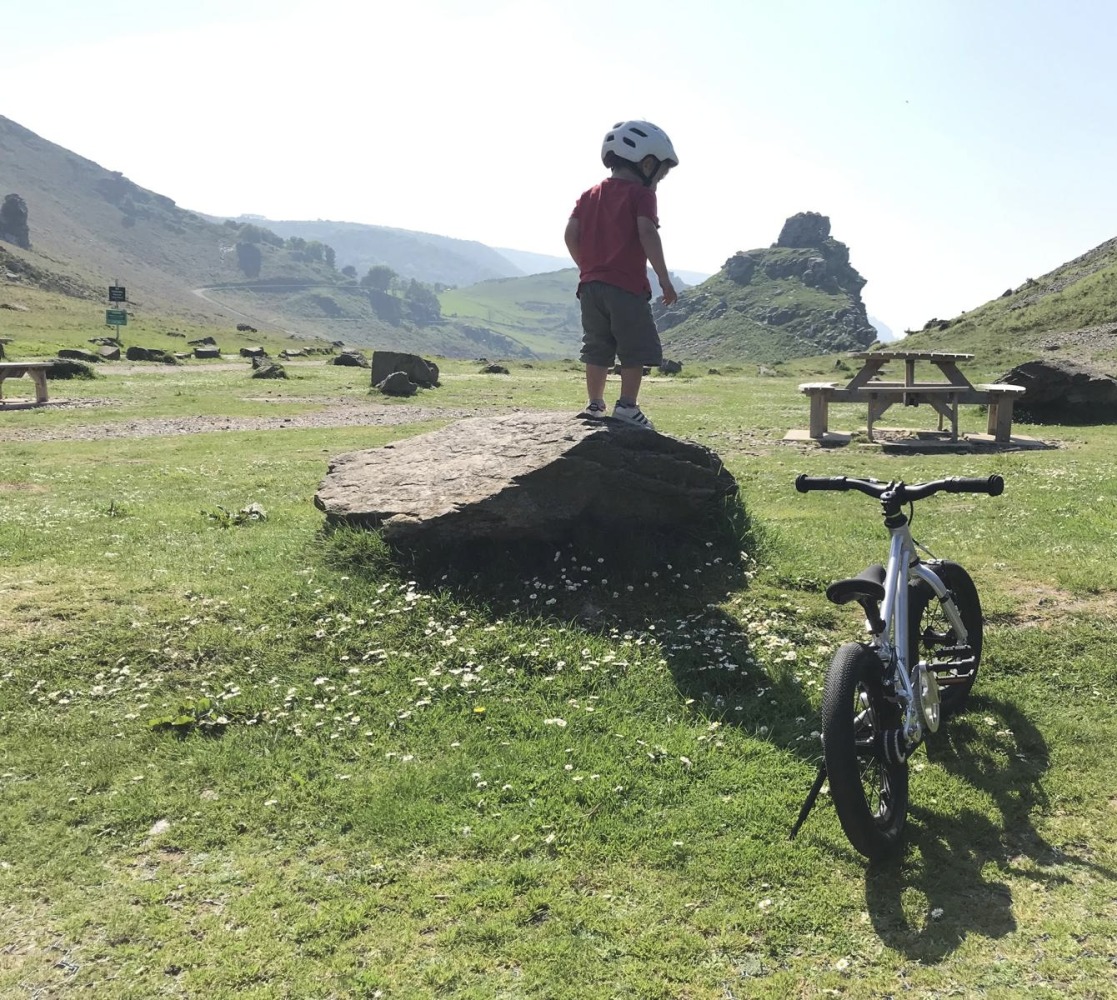 A little boy riding the Early Rider Belter 14 on a sunny day, his bike is propped up on the kickstand and he is standing on a large rock