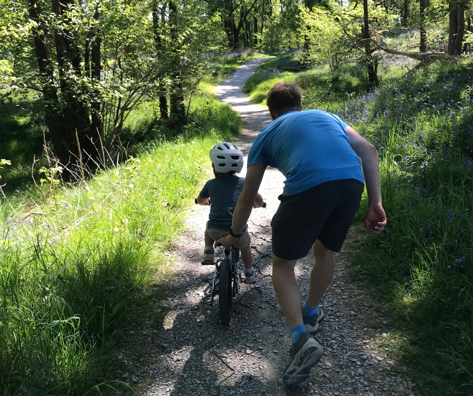 A little boy riding the Early Rider Belter 14 on a sunny day- his dad is pushing him off