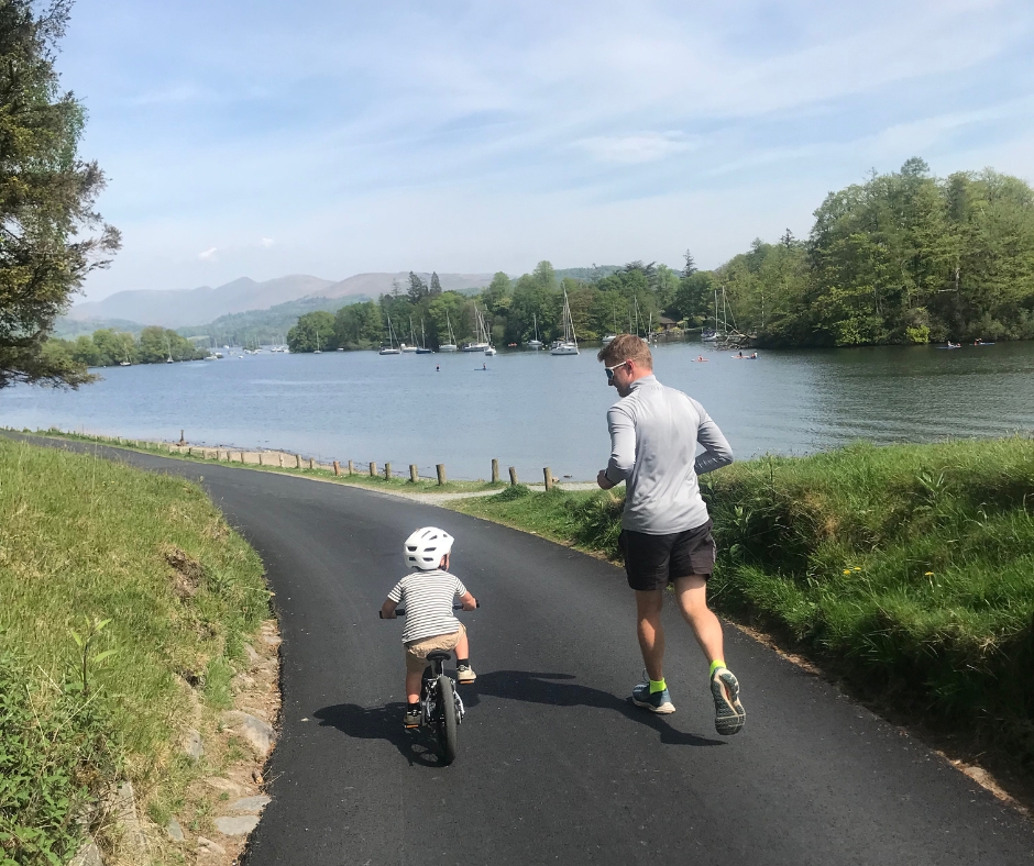 A little boy riding the Early Rider Belter 14 on a sunny day with his dad running alongside next to beautiful views of lake Windermere