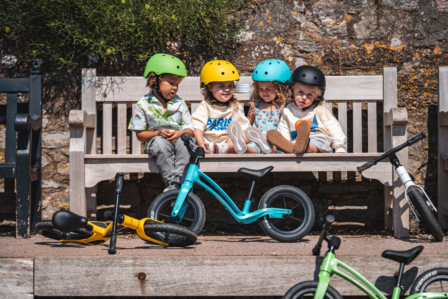 Four toddlers sitting on a wooden bench, all wearing different coloured Hornit AIRO helmets, with their matching AIRO balance bikes scattered before them
