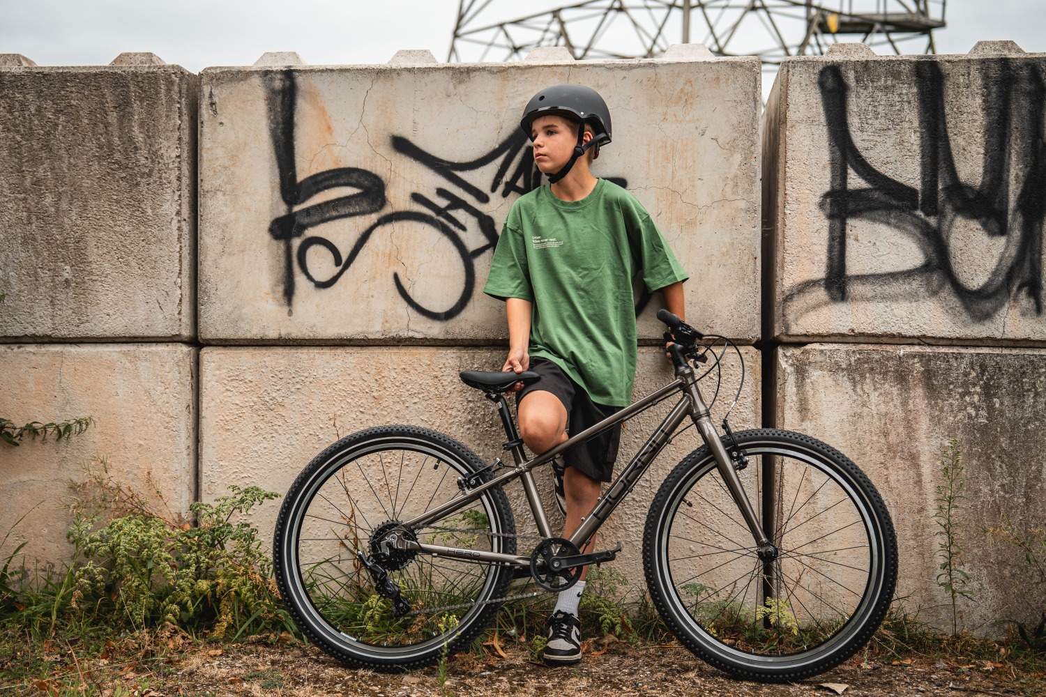 A boy leaning against a wall with one foot resting against it, and a Hornit HERO 24 bike in front of him