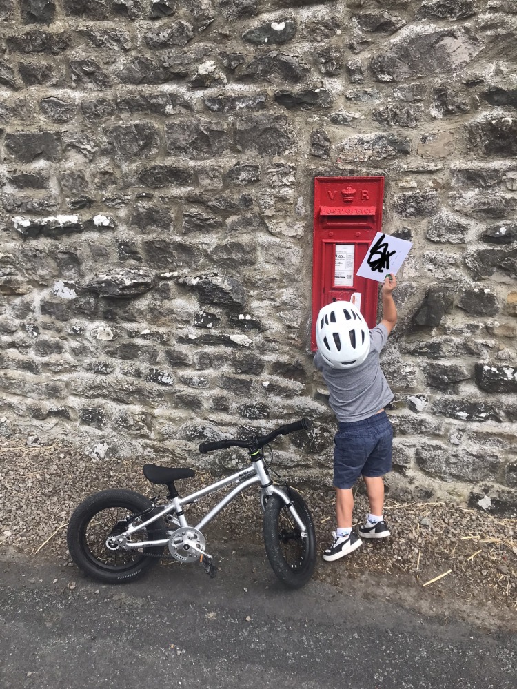 A little boy riding the Early Rider Belter 14 on a sunny day, stopped and putting a letter in a red letterbox