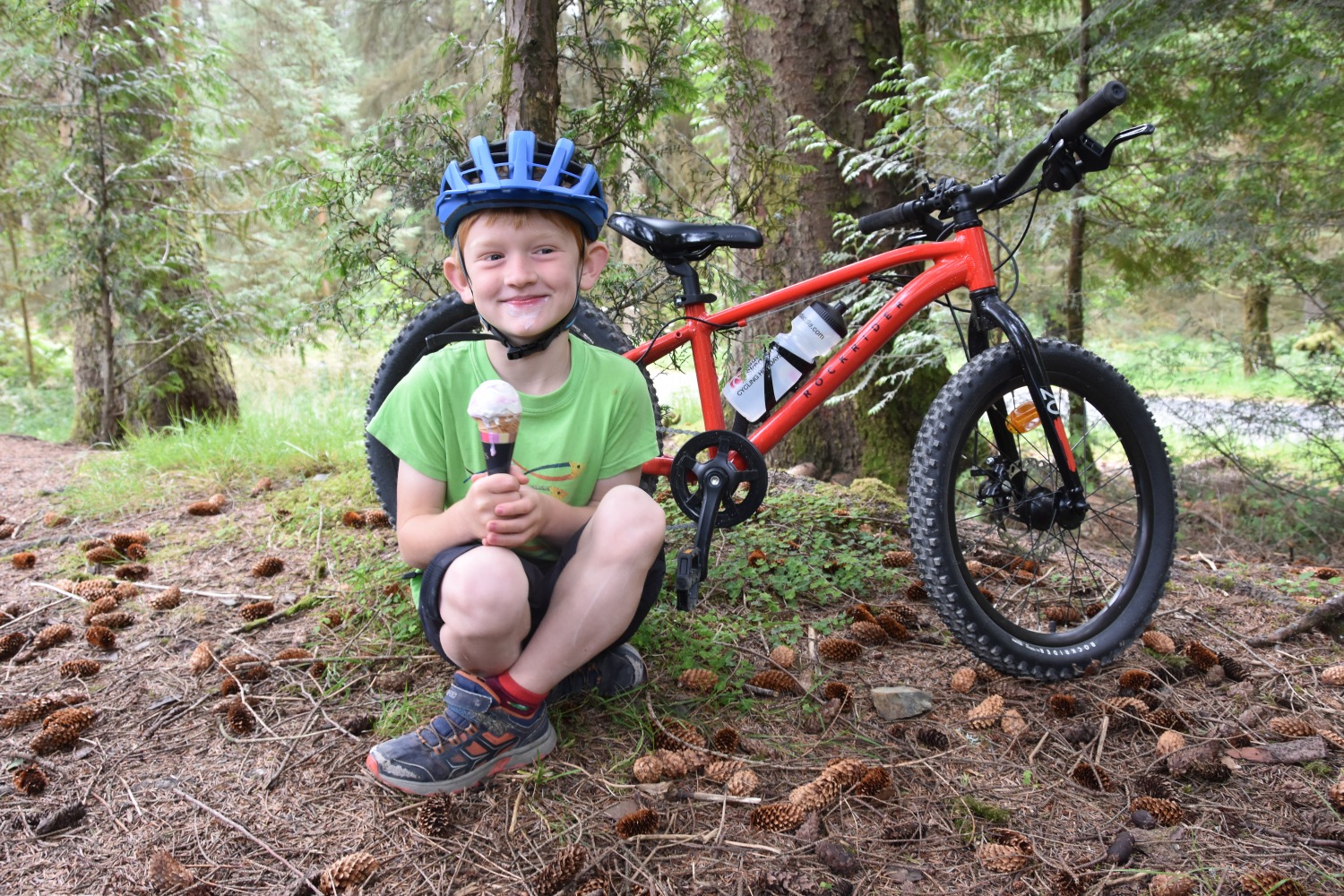 A little boy in a green top eating an ice cream mid ride with his Red 20 inch BTWIN EXPL 900r behind him