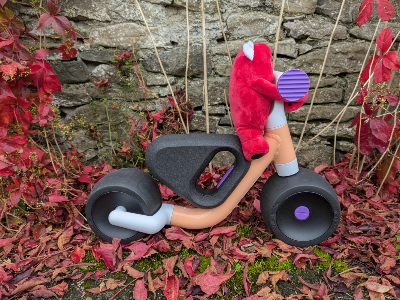 the peach colour woom WOW toddler first ride-on bike pictured against a wall with red leaves on the ground