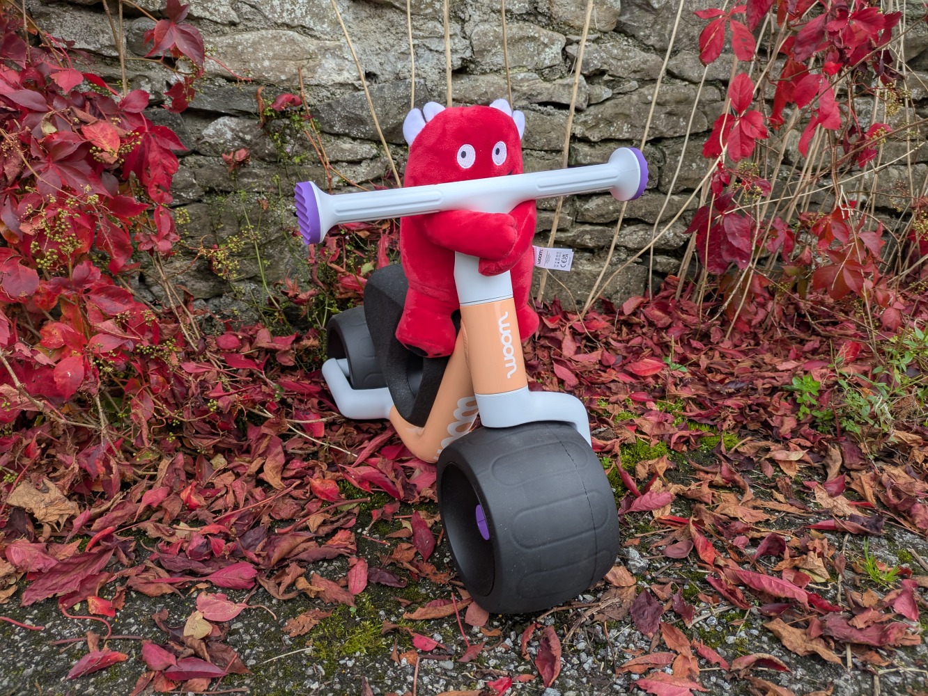 the peach colour woom WOW toddler first ride-on bike pictured against a wall with red leaves on the ground