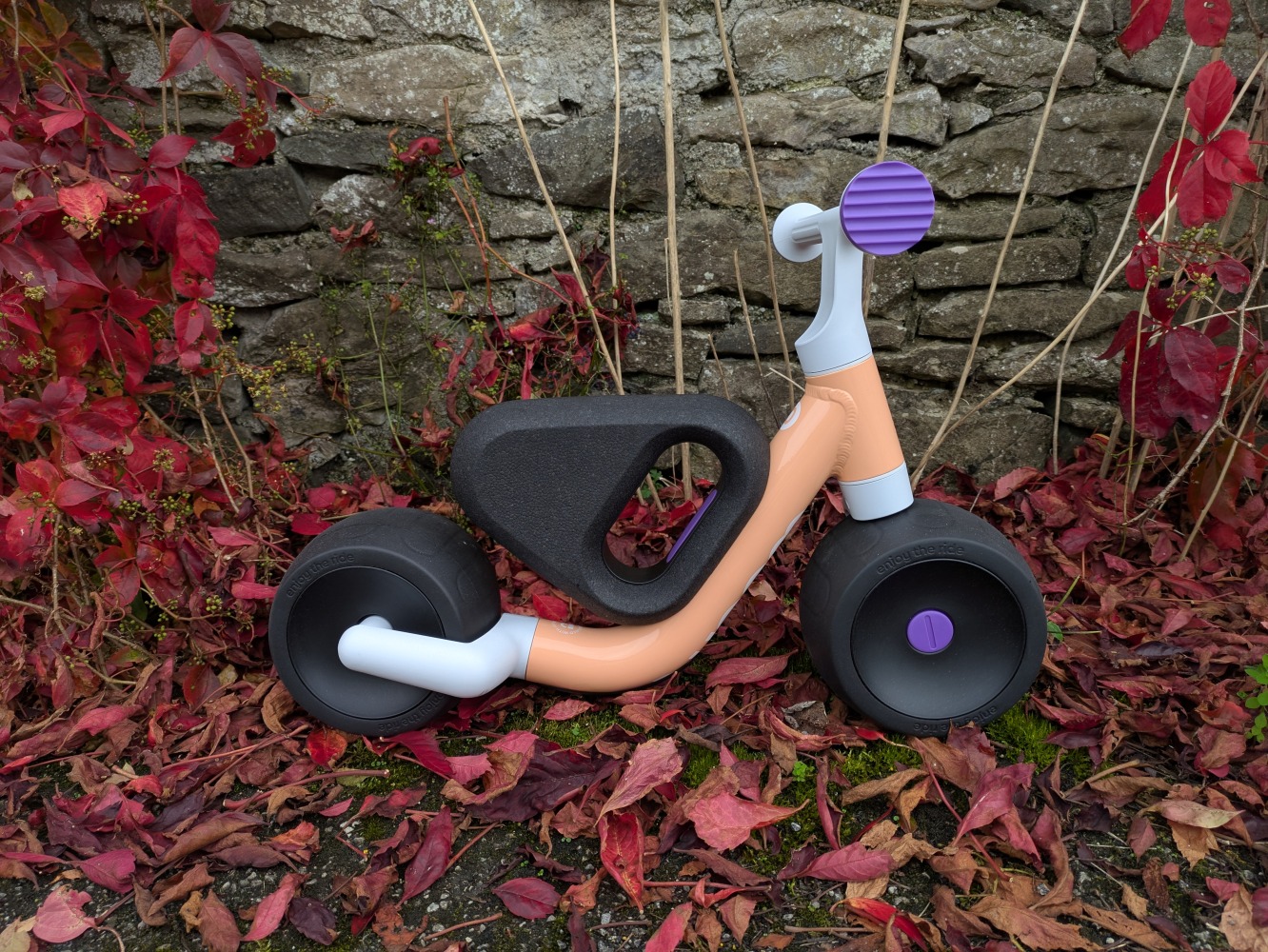 the peach colour woom WOW toddler first ride-on bike pictured against a wall with red leaves on the ground