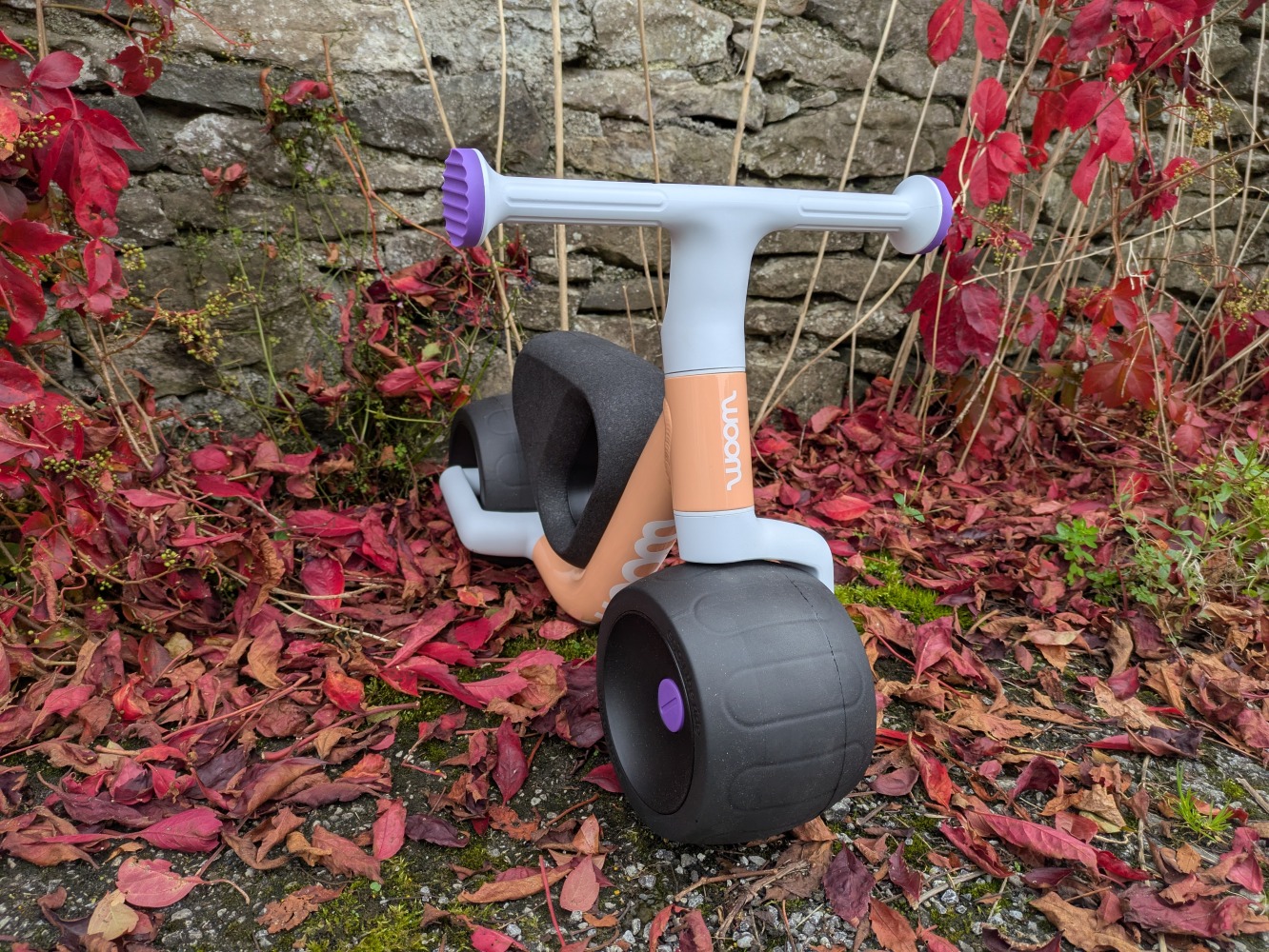the peach colour woom WOW toddler first ride-on bike pictured against a wall with red leaves on the ground