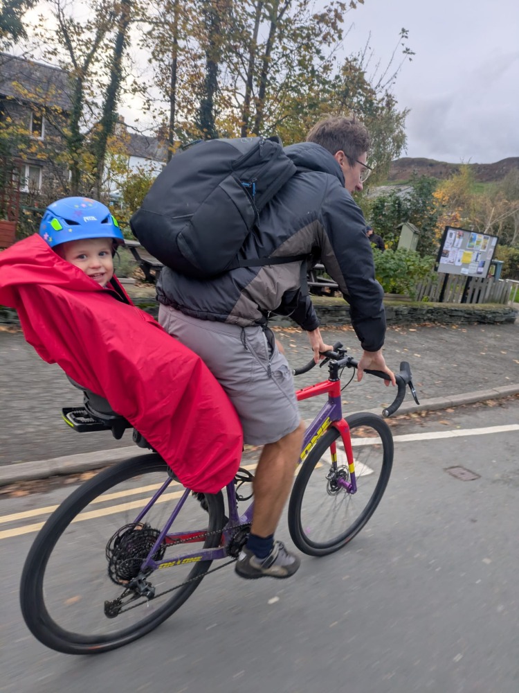 a little boy on a rear bike seat in his red Hamax Bike Seat Rain Poncho on his dads bike