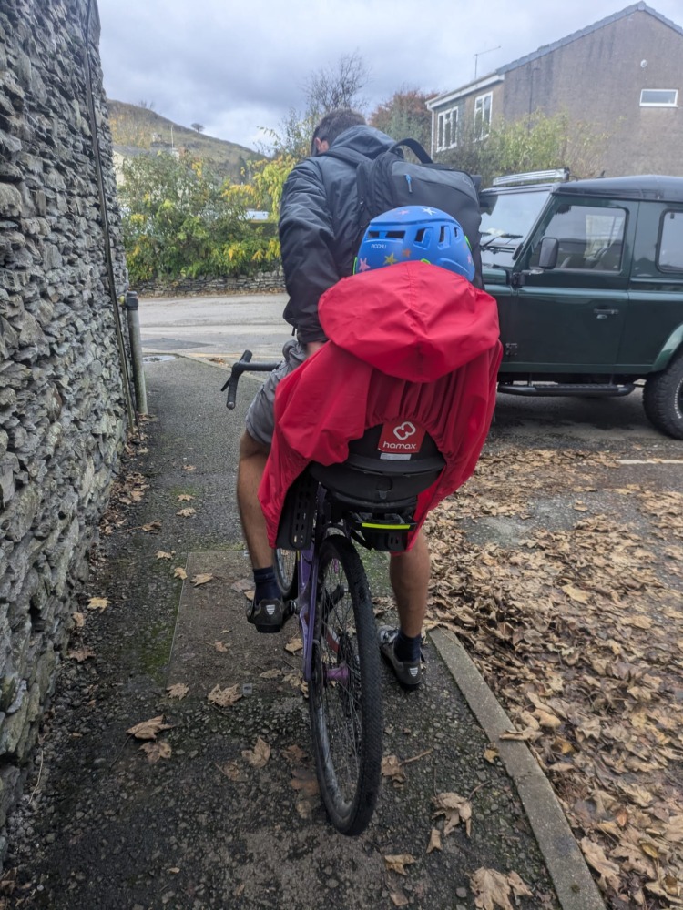 a little boy on a rear bike seat in his red Hamax Bike Seat Rain Poncho on his dads bike