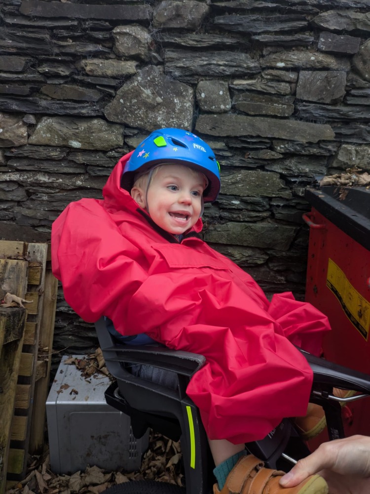 a little boy on a rear bike seat in his red Hamax Bike Seat Rain Poncho smiling