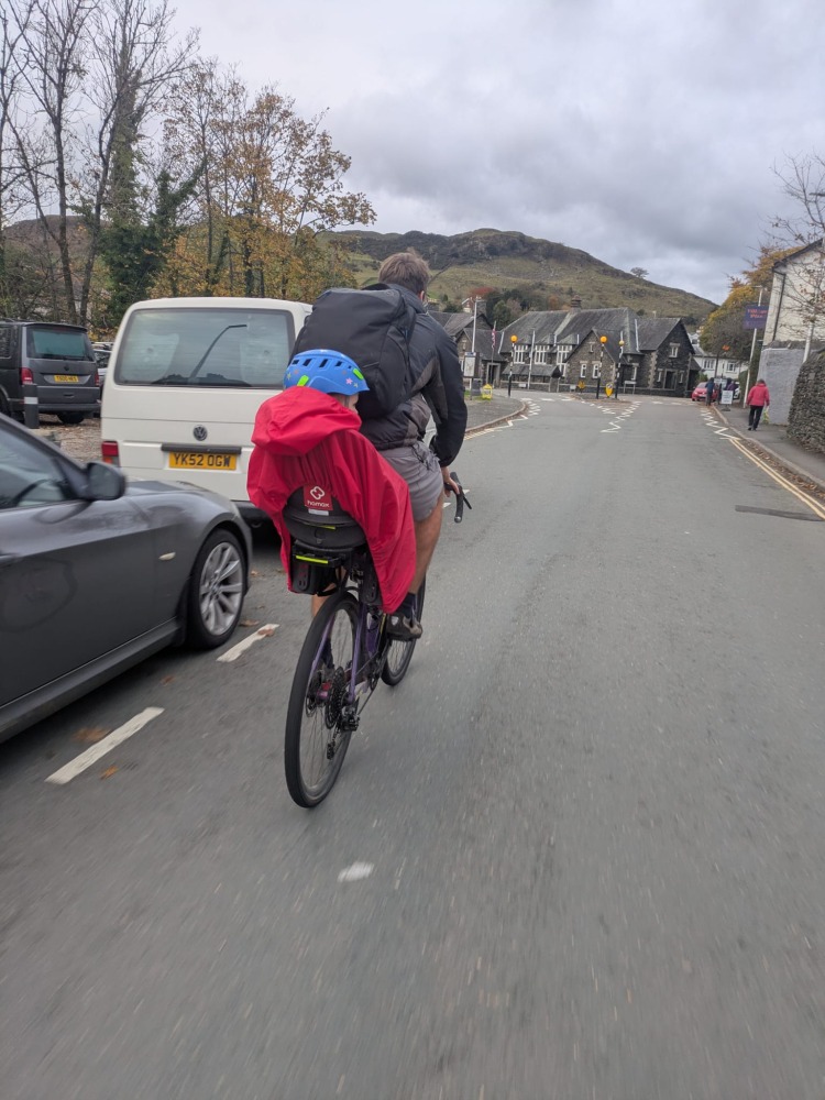 a little boy on a rear bike seat in his red Hamax Bike Seat Rain Poncho on his dads bike