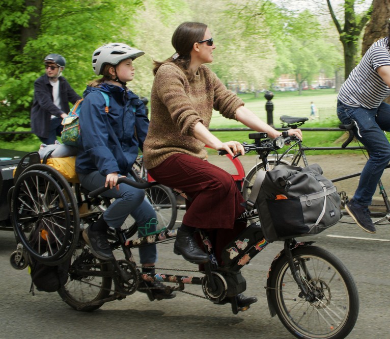 Kid3 and me riding our e-tandem through a park with other cyclists on a Kidical Mass ride. My wheelchair is on the back of the tandem and there’s a cuddly grey toy cat tied to the wheelchair, too.