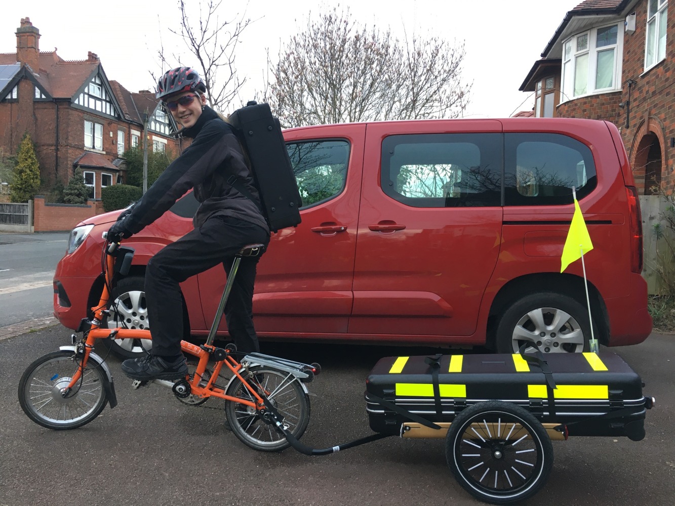 Kid1 is in front of a red van. He is on an orange e-converted Brompton, smiling at the camera. He has a big black rectangular rucksack on his back containing an alto saxophone, and is towing a trailer with a very large black baritone saxophone case tied to it. The case has high-vis yellow bands stuck to it, and a yellow flag stuck on the rear right.