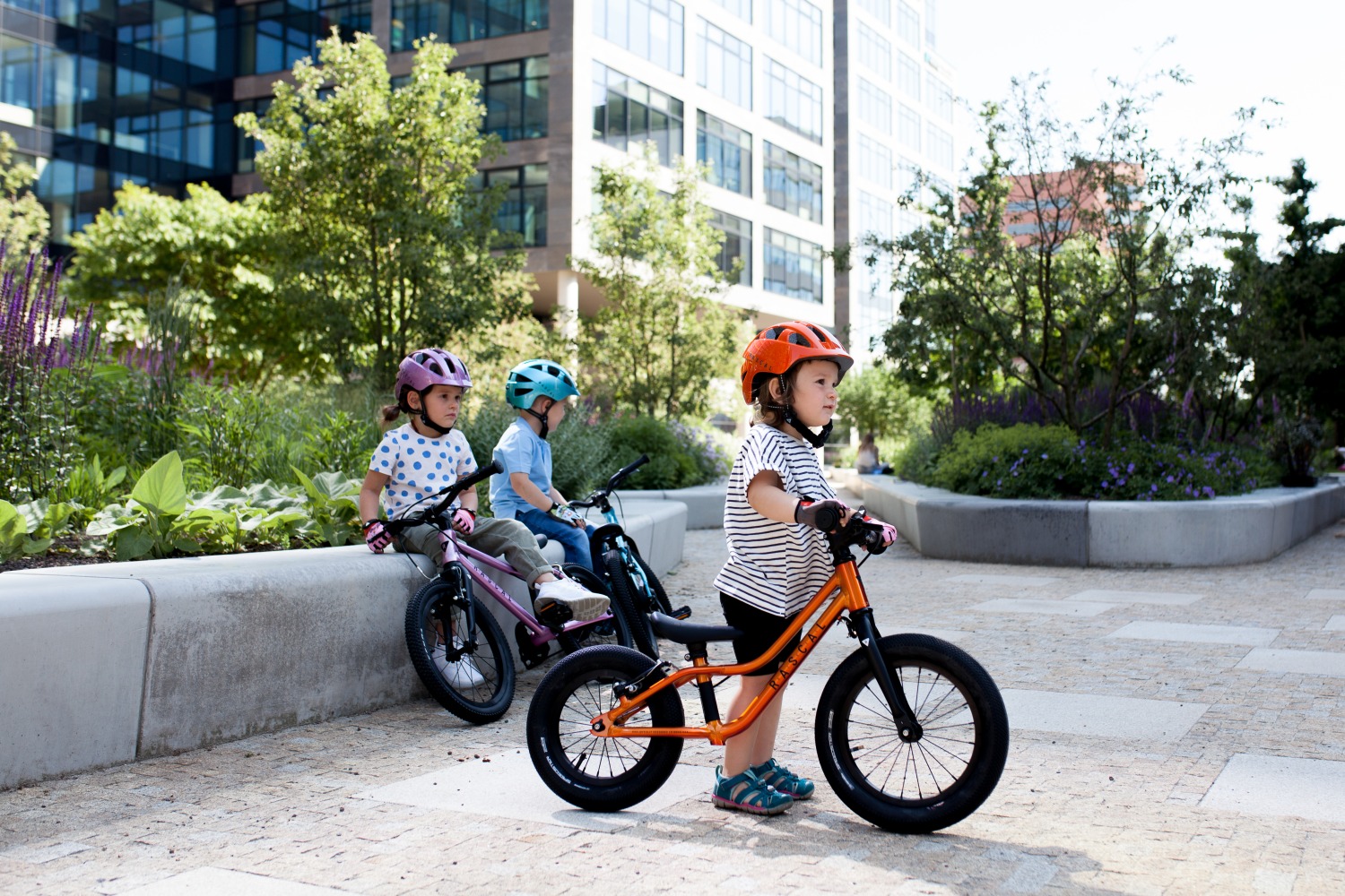 Three children of different ages on Rascal bikes,  The youngest is on a balance bike and the other two are on pedal bikes.  They are in a traffic free urban area with white paving slabs, with offices in the background